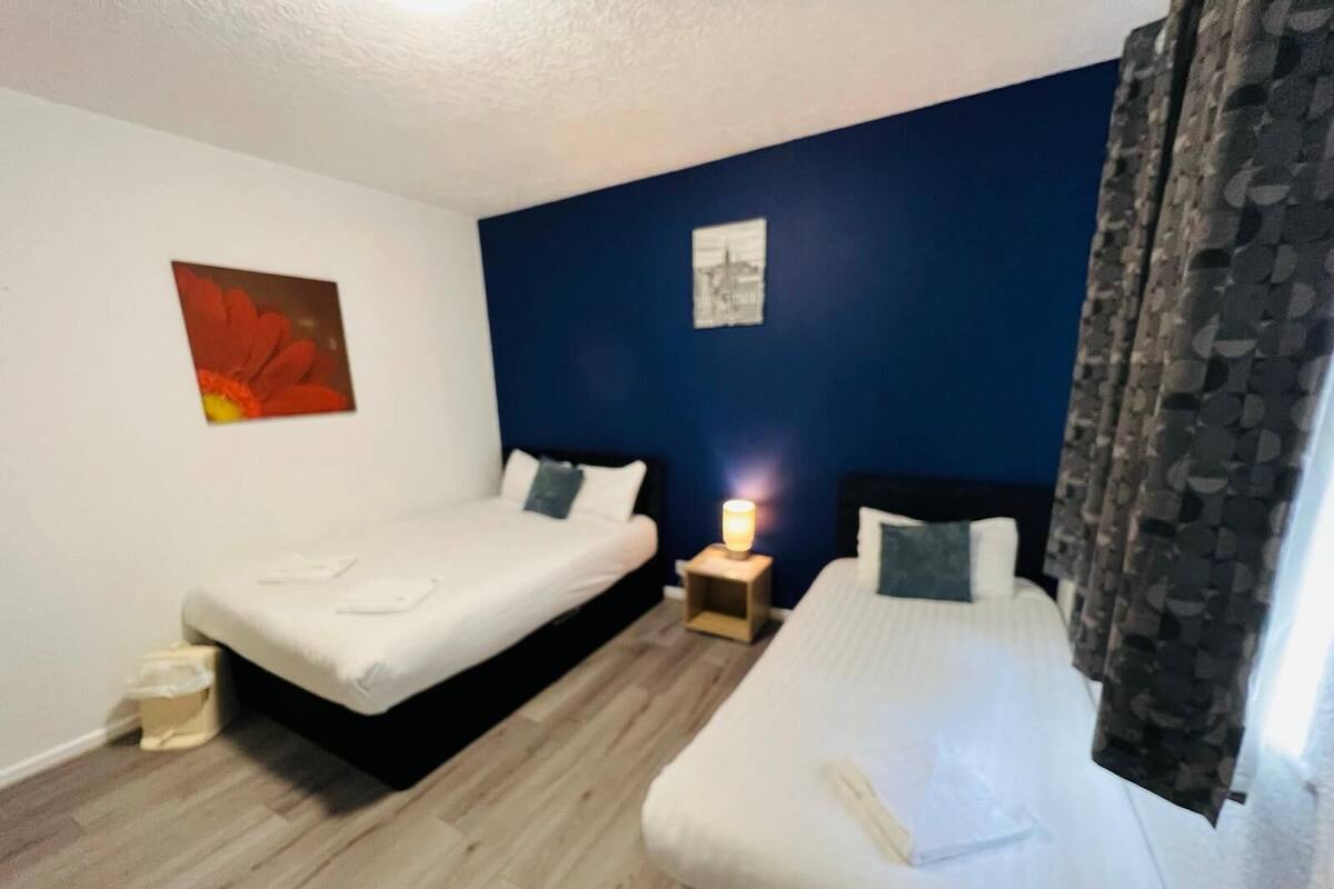 A hotel room with two beds, a nightstand with a lamp, a small trash bin, and a painting on the wall.