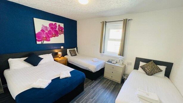 A hotel room with two beds, a nightstand with a lamp, a small table with a kettle, and a window with curtains. - Calypso Hotel Blackpool (Blackpool)