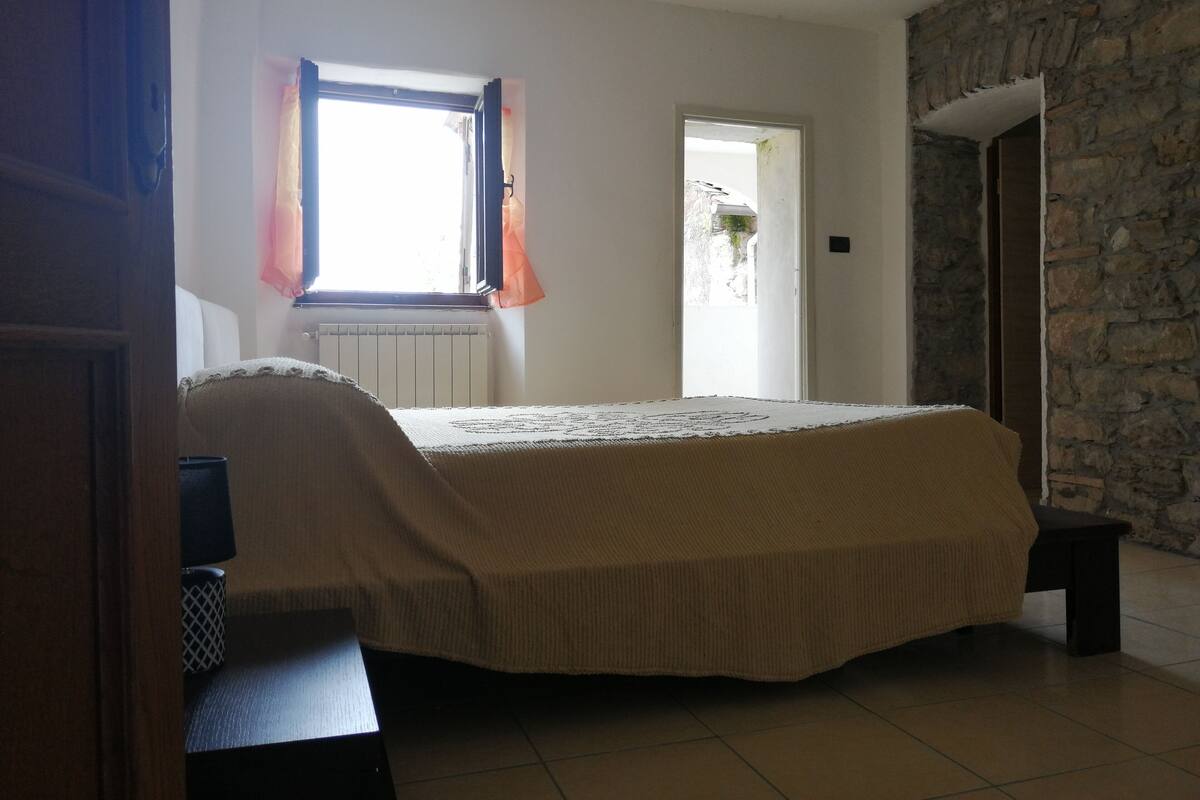 A bedroom with a bed, a radiator, a window with curtains, and a stone wall.