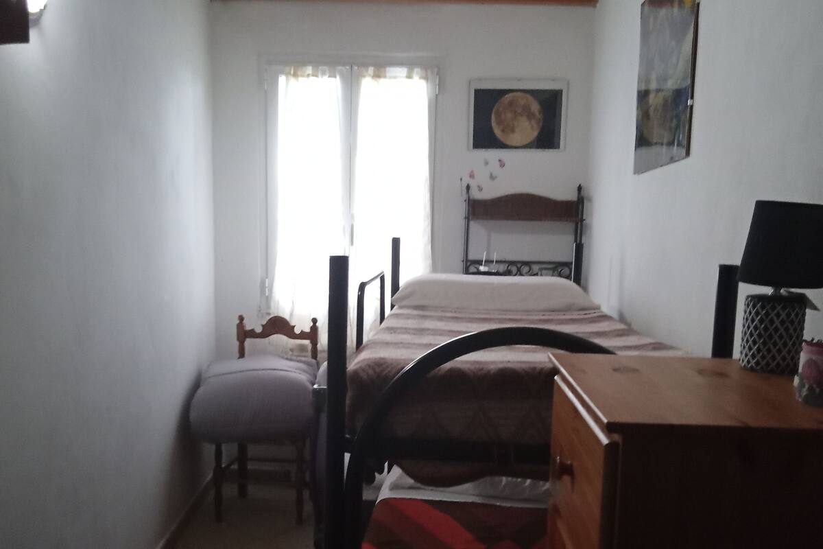 A small, dimly lit room with a bed, a chair, a dresser, and a lamp.