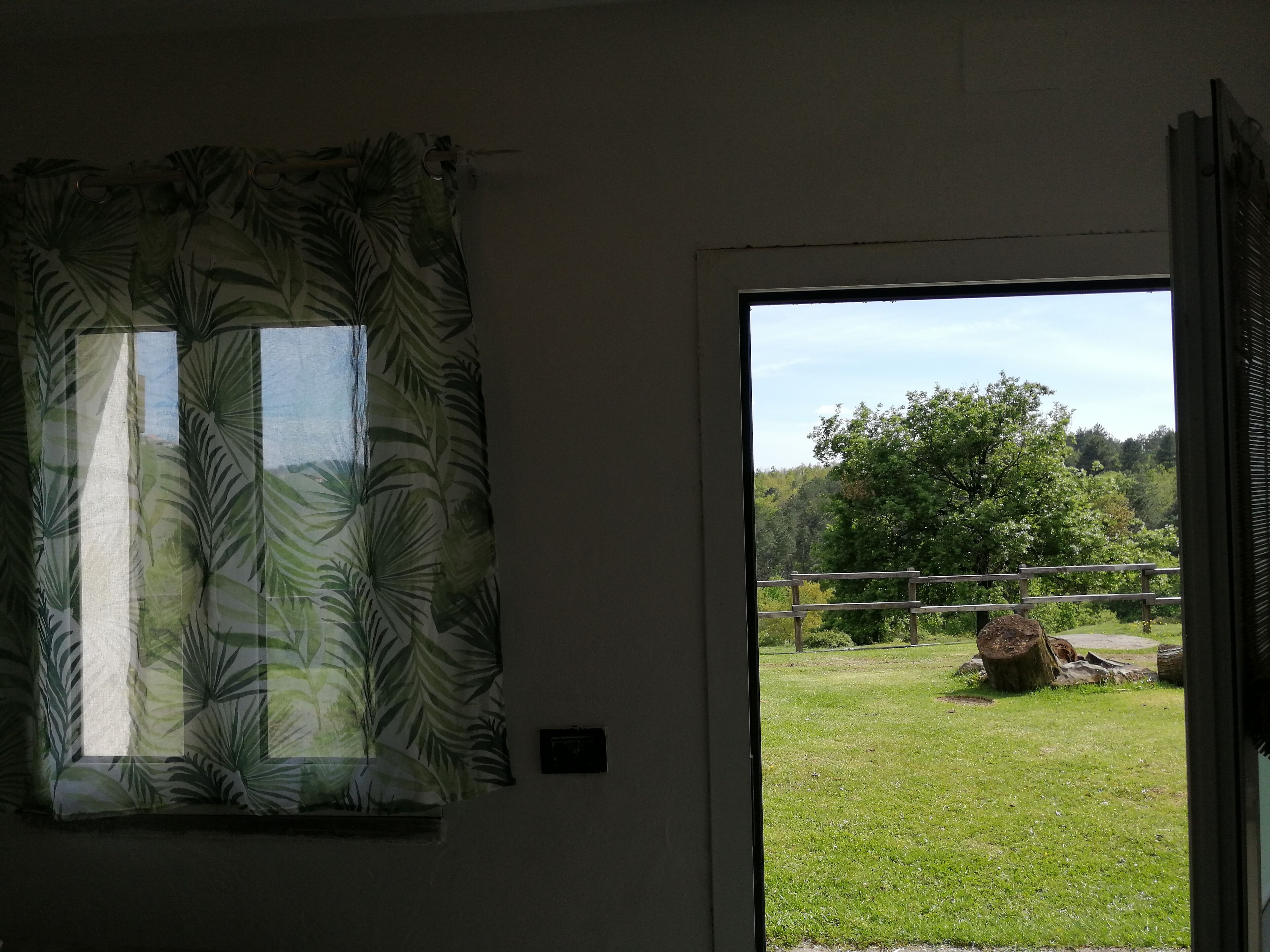 A room with a window featuring a tropical leaf pattern curtain, a view of a grassy outdoor area with a wooden fence and trees.