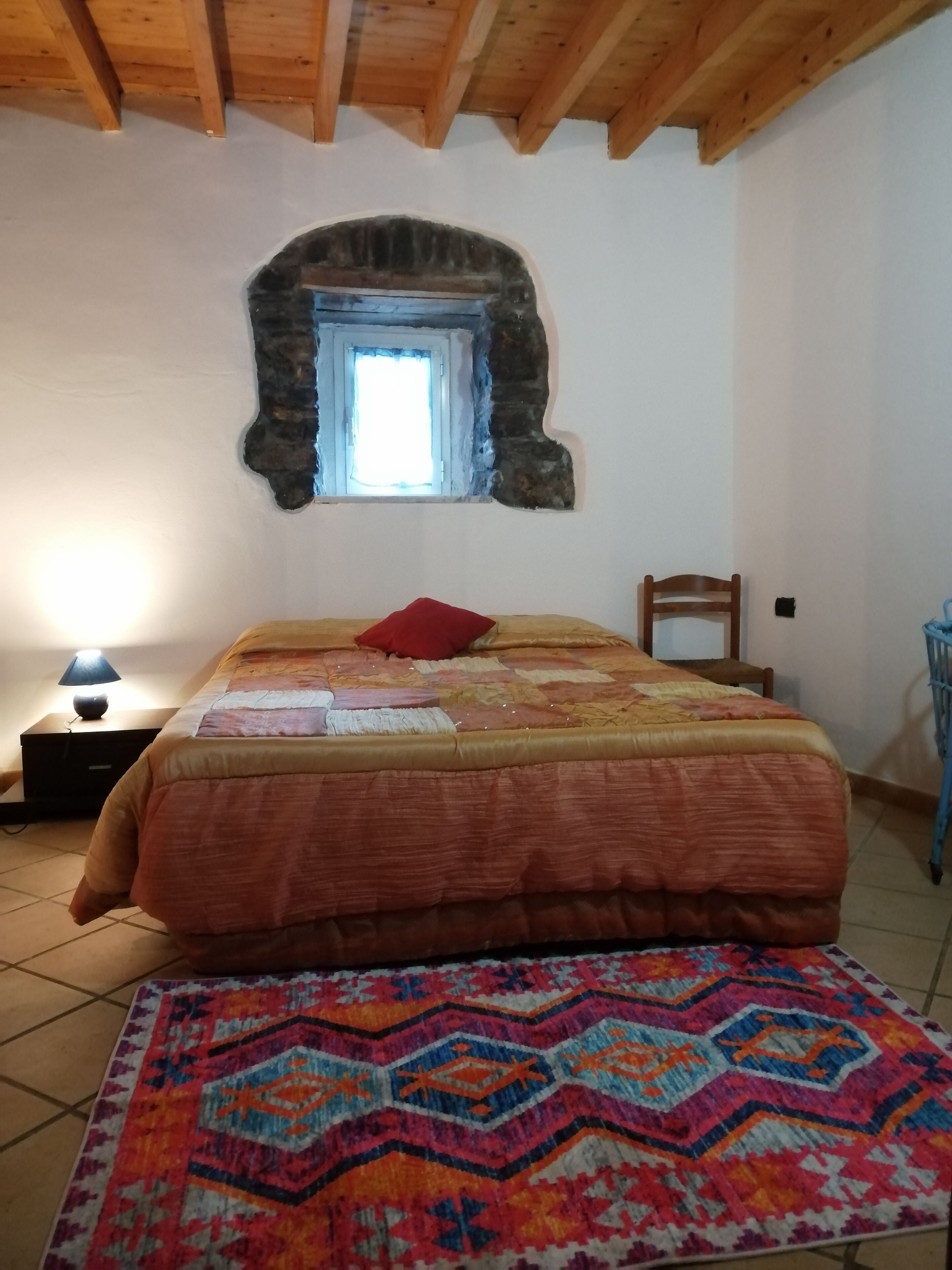 Quarto com cama, tapete, mesa de cabeceira, luminária, cadeira e parede de pedra com janela.