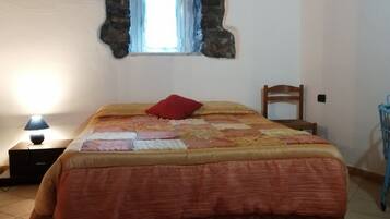 Quarto com cama, tapete, mesa de cabeceira, luminária, cadeira e parede de pedra com janela.