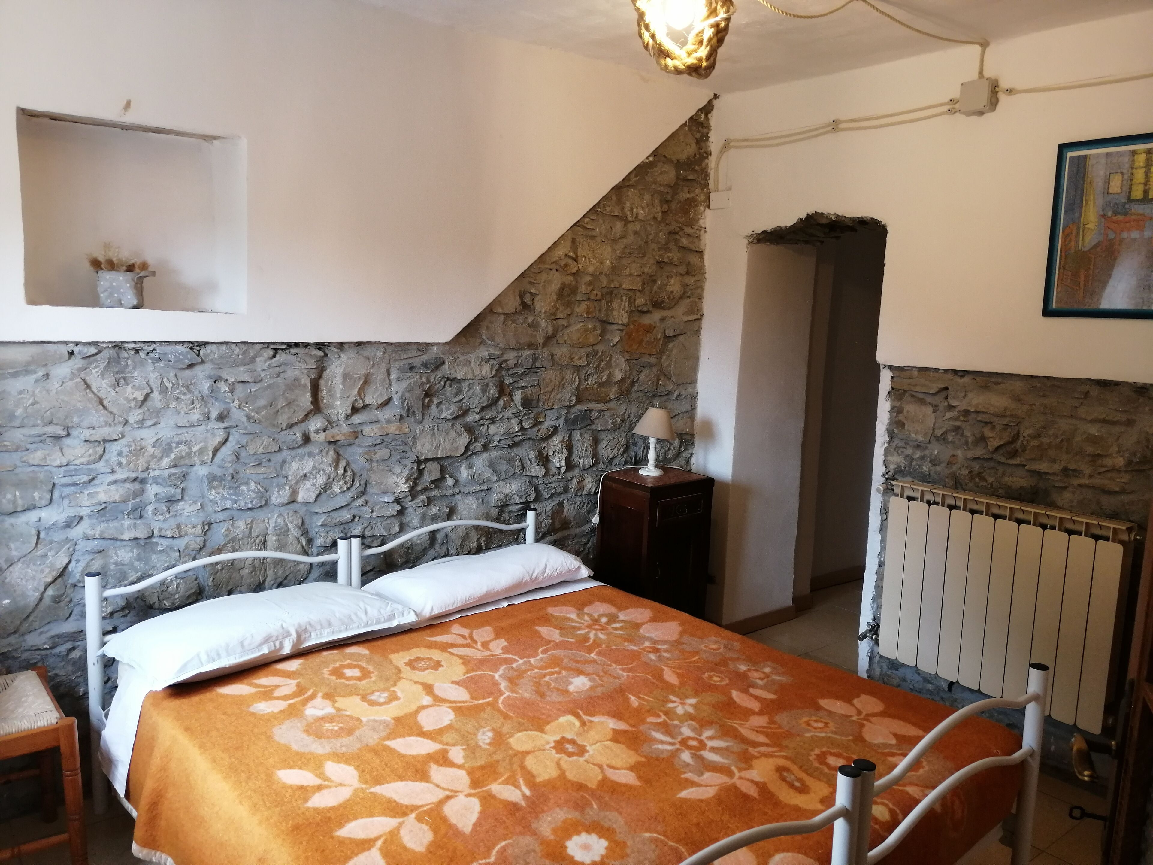 Un dormitorio con pared de piedra, cama con colcha floral, mesita de noche con lámpara y un radiador.