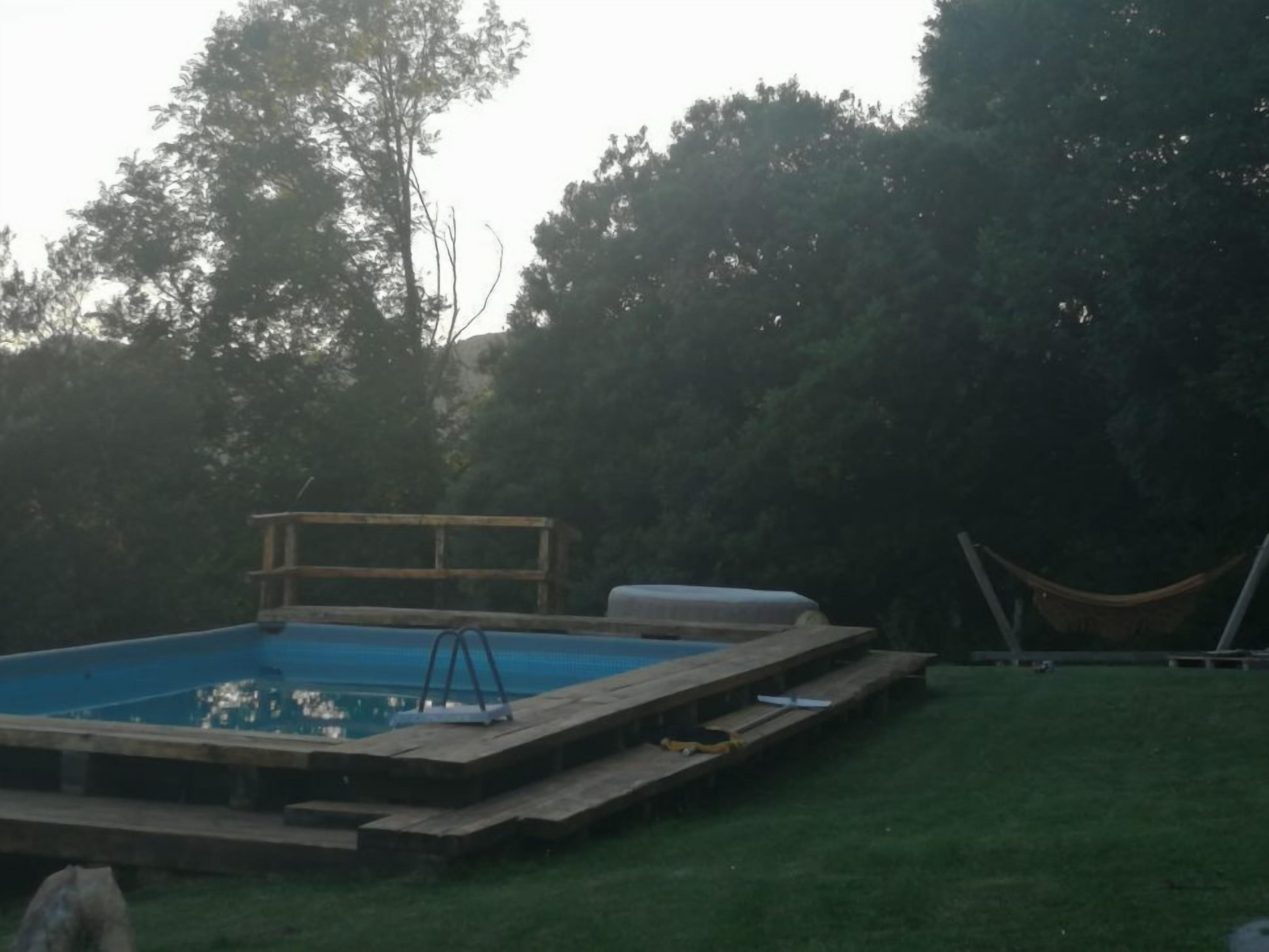 A rectangular swimming pool with a wooden deck, surrounded by a grassy yard and trees.
