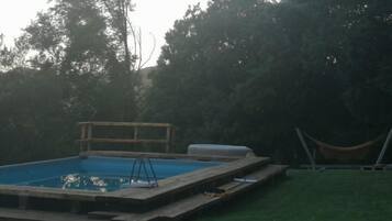 A rectangular swimming pool with a wooden deck, surrounded by a grassy yard and trees.