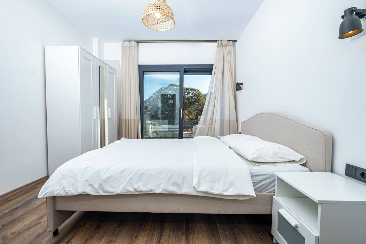 A modern bedroom with a large bed, a bedside table, a lamp, and a view of the outdoors through a sliding glass door.