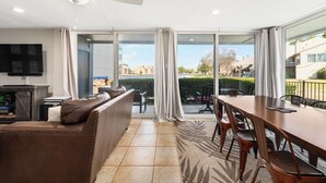 A modern living room with a sofa, a dining table, and chairs. There is a television and a speaker on a stand. The room has large windows with curtains and a view of buildings and a body of water outside. - Lakefront Escape with a View! Your Lake Conroe Vacation Starts Here (Montgomery)