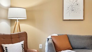 Ein Wohnzimmer mit einem braunen Ledersessel, einem grauen Sofa, einer weißen Lampe und einem gerahmten Blumenbild an der Wand.