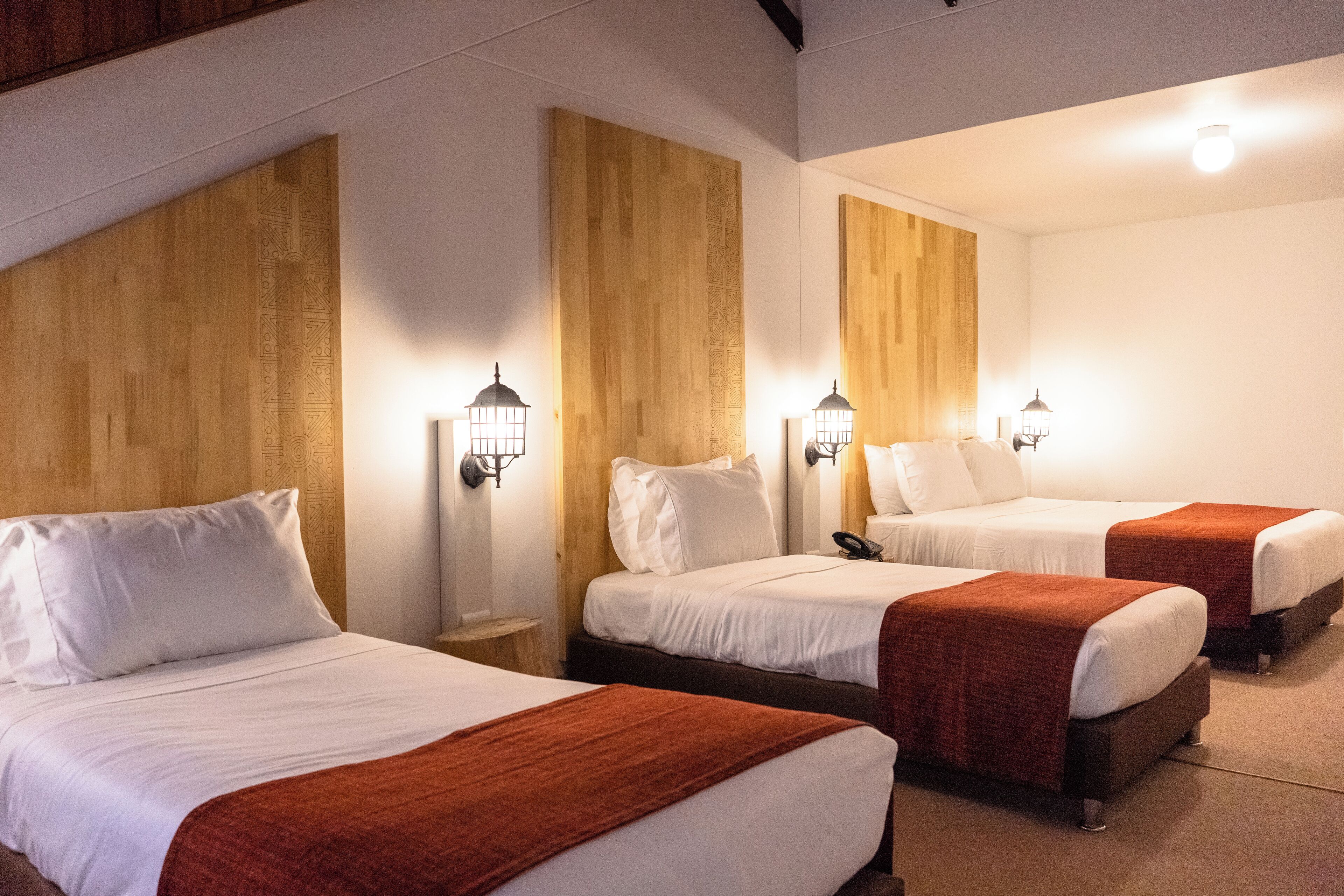 A hotel room with three beds, each with white bedding and a brown blanket. There are wall-mounted lamps on either side of the beds.
