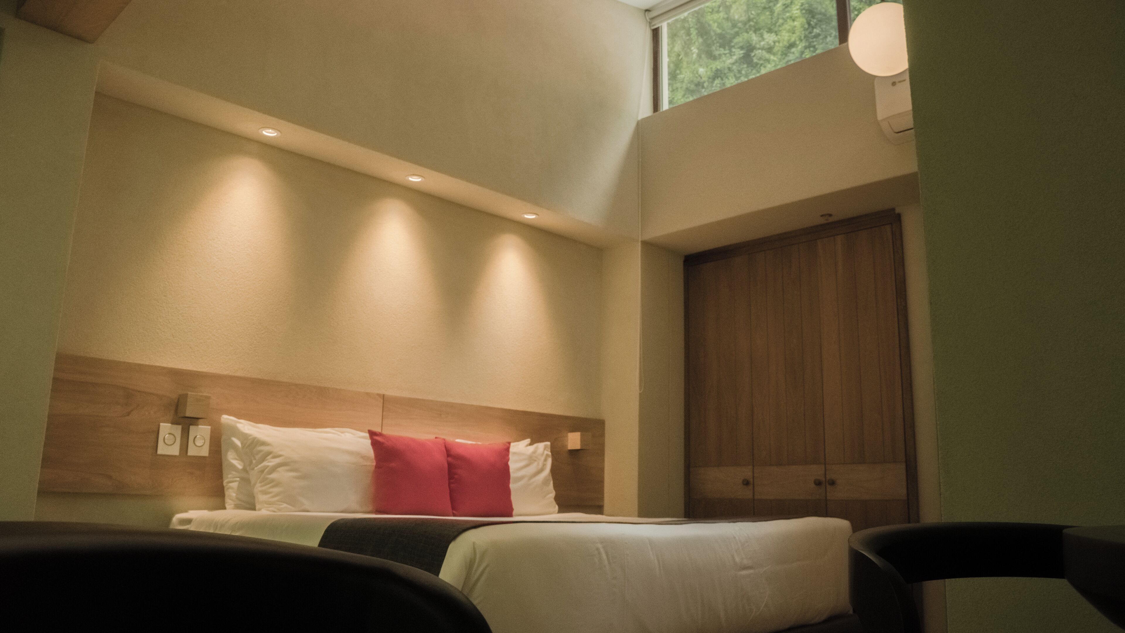 A hotel room with a bed, a wooden wardrobe, and a window with a view of greenery.
