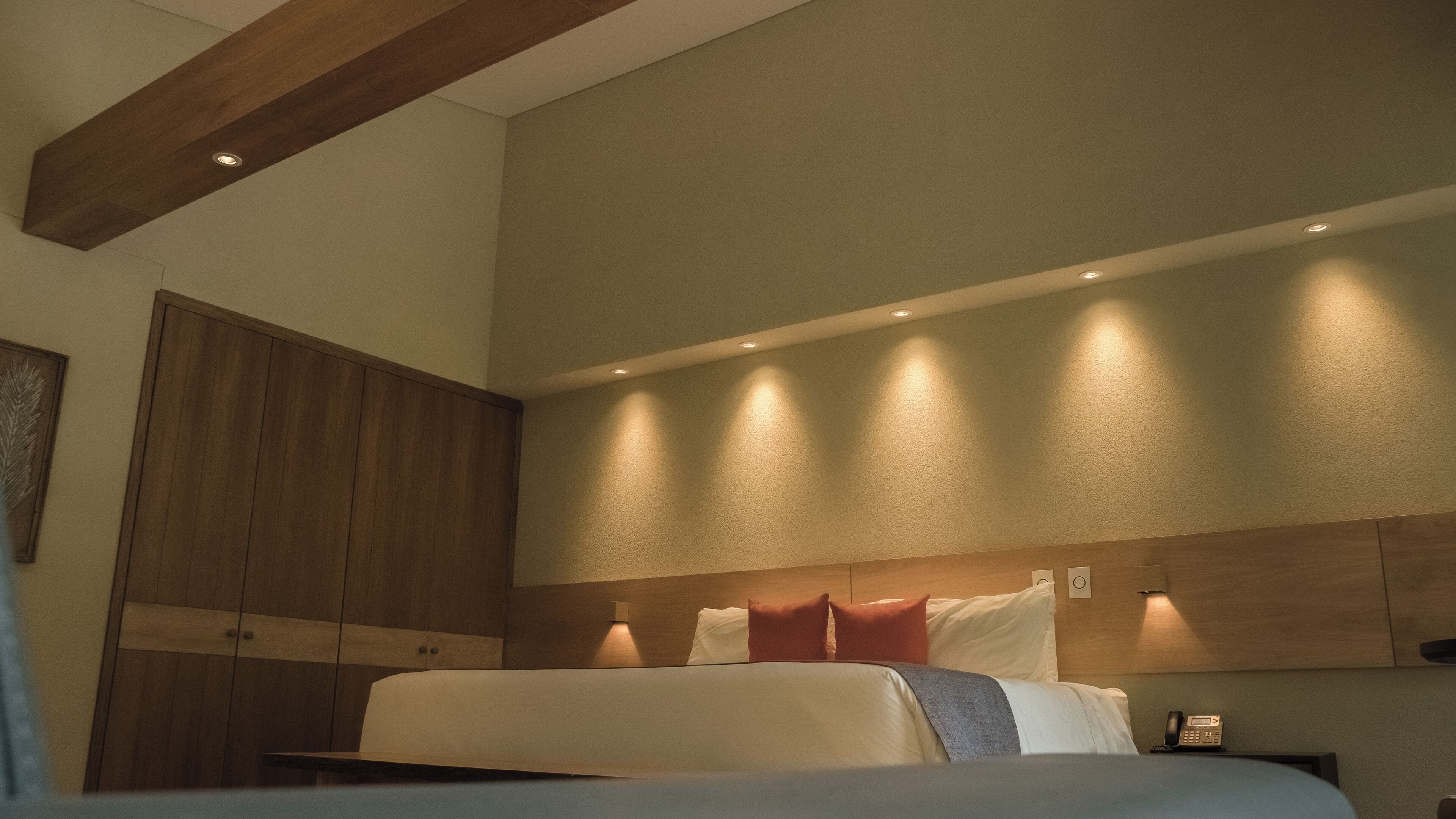 A bedroom with a bed, wooden wardrobe, recessed ceiling lights, and a bedside table with a phone.