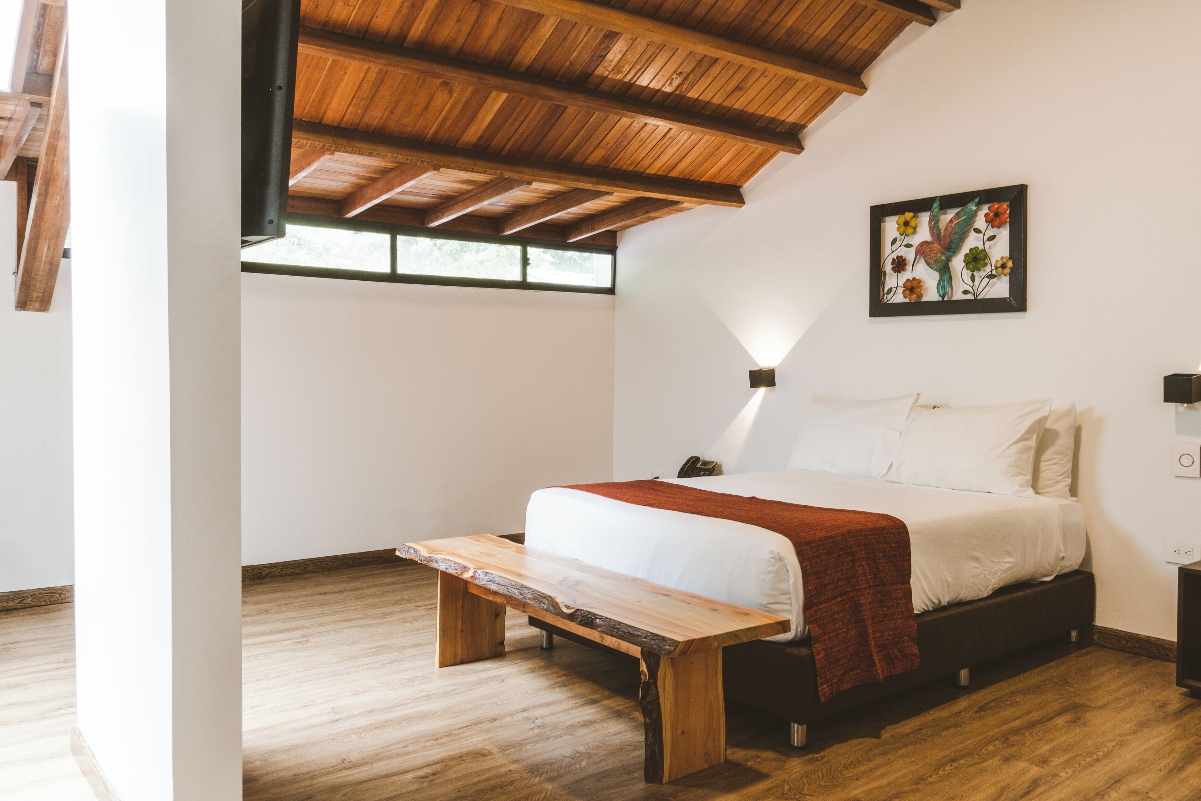 Une chambre avec un plafond en bois, un lit avec une literie blanche, un banc et un luminaire fixé au mur.