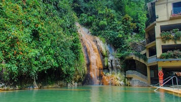 Une cascade se jetant dans un bassin, entouré d’une végétation luxuriante, et un bâtiment doté de balcons.