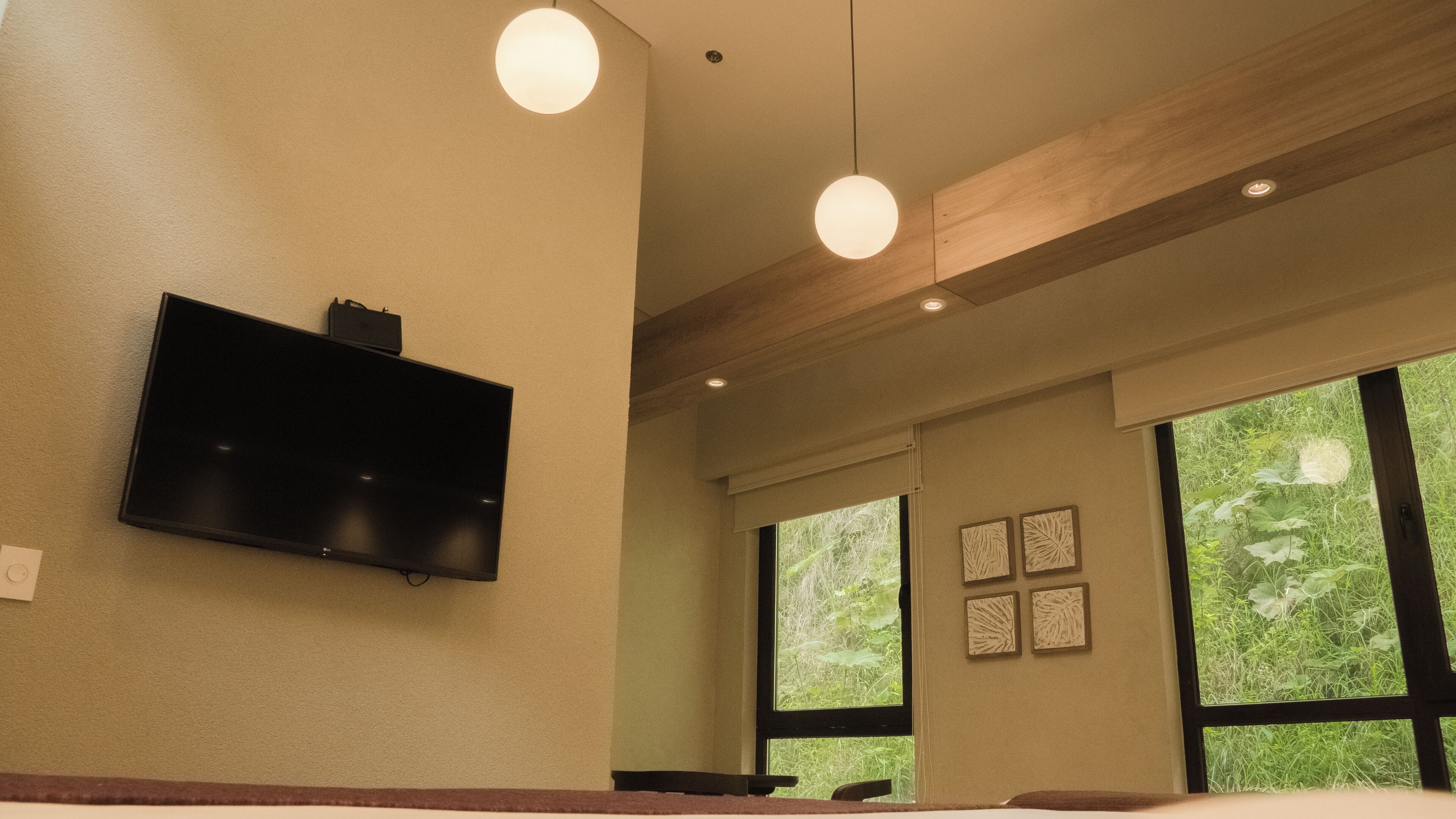 A modern hotel room with a flat-screen TV mounted on the wall, large windows with a view of greenery, and a wooden ceiling beam.