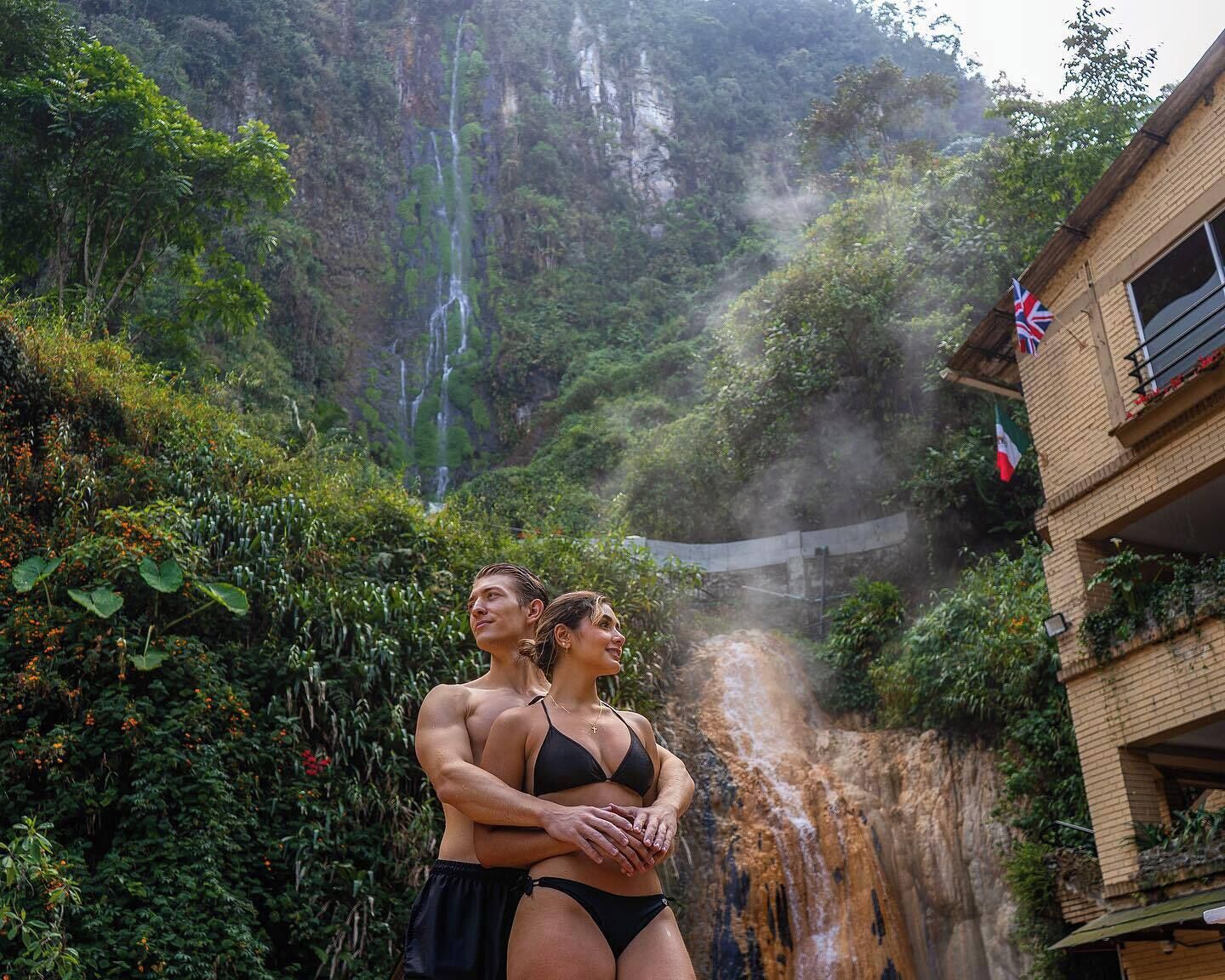 Una pareja frente a una cascada, con abundante vegetación y un edificio con balcón.