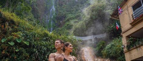 Una pareja frente a una cascada, con abundante vegetación y un edificio con balcón.