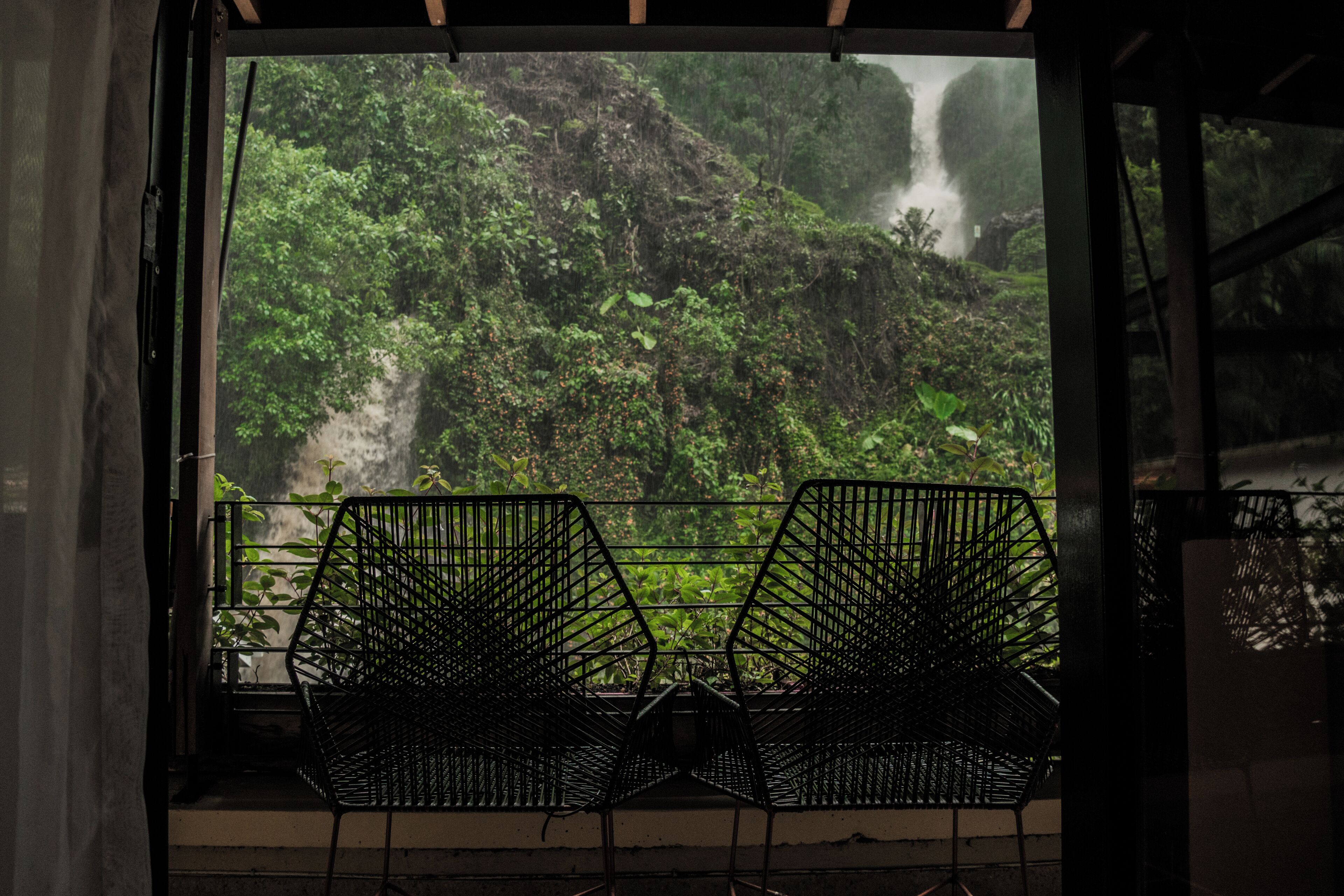 Une pièce avec une fenêtre donnant sur une forêt luxuriante et une cascade. Deux chaises en métal noir sont placées devant la fenêtre.