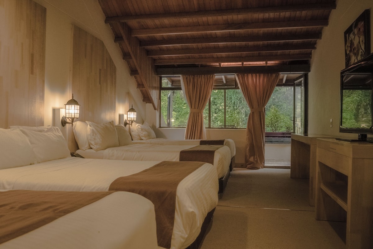 Ein Hotelzimmer mit drei Betten, Holzbalken, einem Schreibtisch mit Fernseher und Fenstern mit Vorhängen.