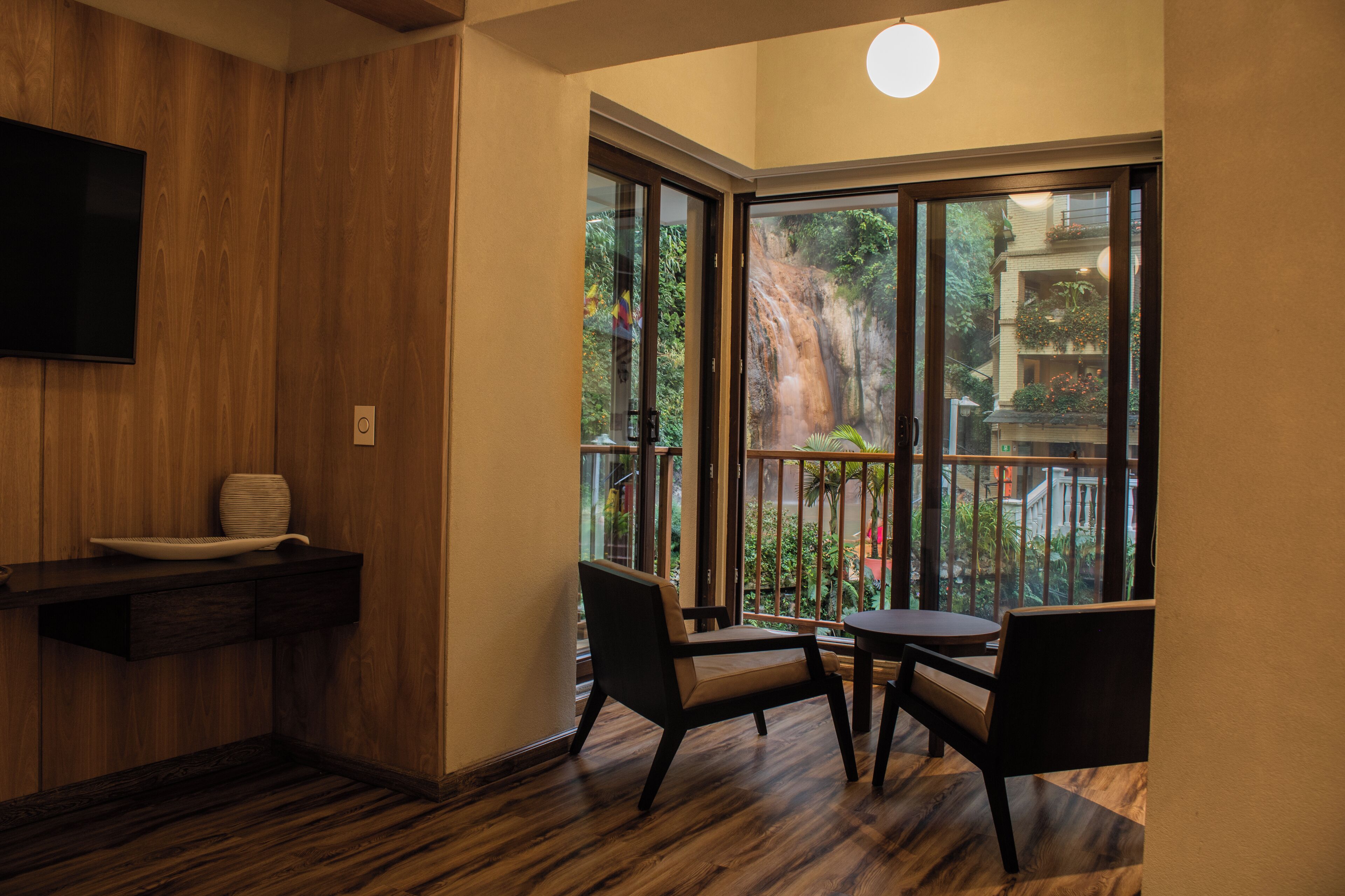 Habitación de esquina con piso de madera, televisor de pantalla plana en la pared, una mesita con dos sillas y un ventanal con vistas a un jardín y un edificio.