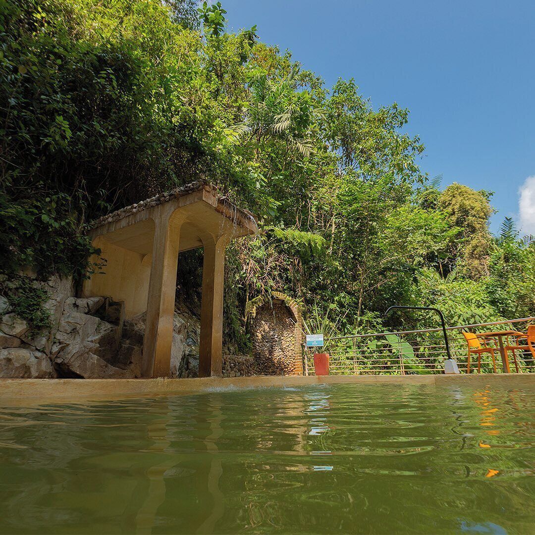 Un pequeño pabellón rústico con base de piedra y techo de tejas, ubicado junto a una masa de agua tranquila rodeada de exuberante vegetación y árboles.