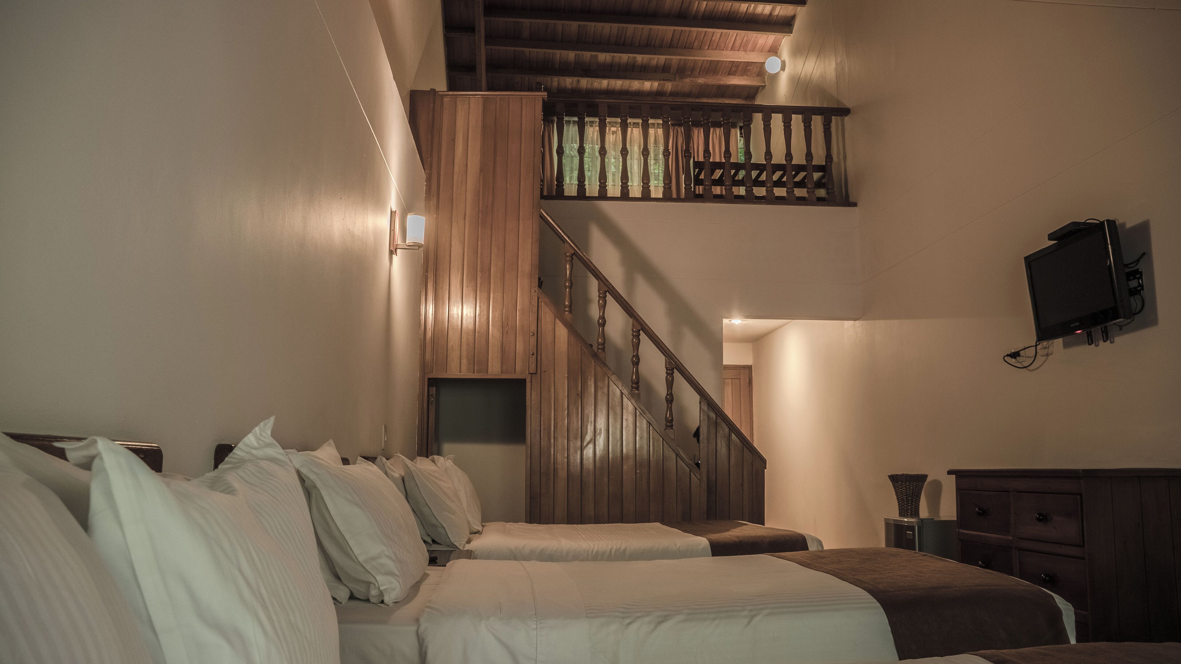 A bedroom with two beds, a wooden staircase, a TV mounted on the wall, and a small table.