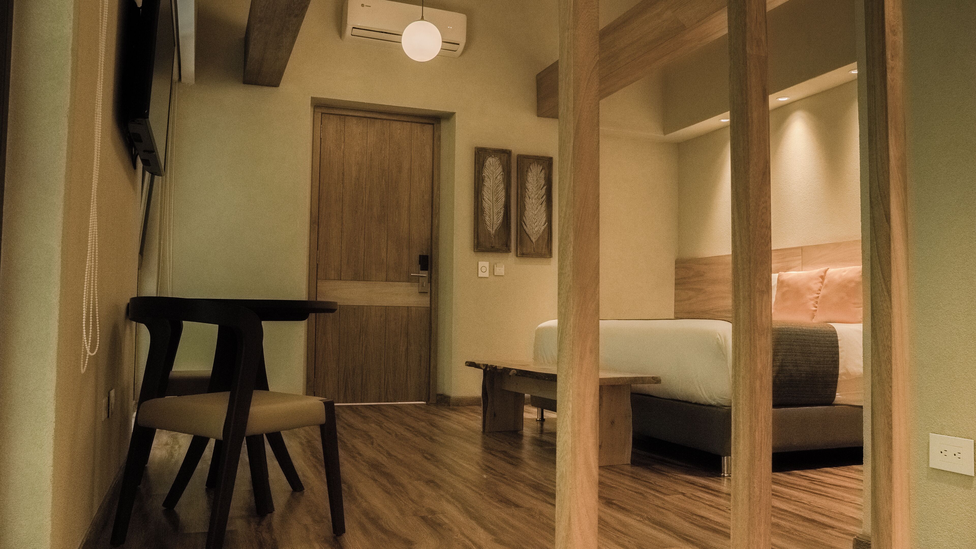 A hotel room with a wooden floor, a bed with a headboard, a small table, a chair, and a wardrobe.