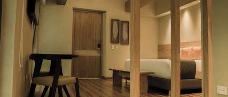 A hotel room with a wooden floor, a bed with a headboard, a small table, a chair, and a wardrobe.