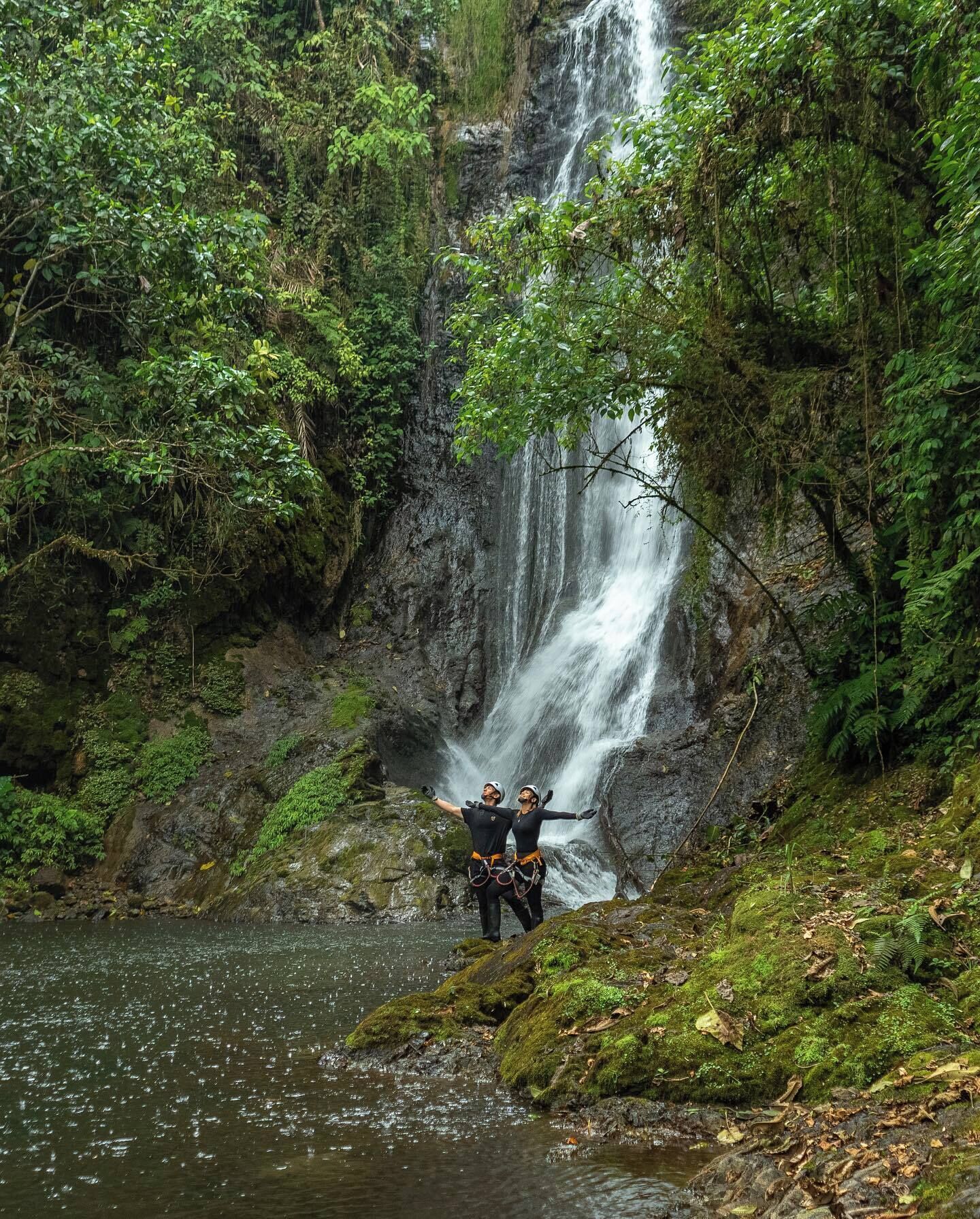 A waterfall in a forested area with two people standing on a moss-covered rock.