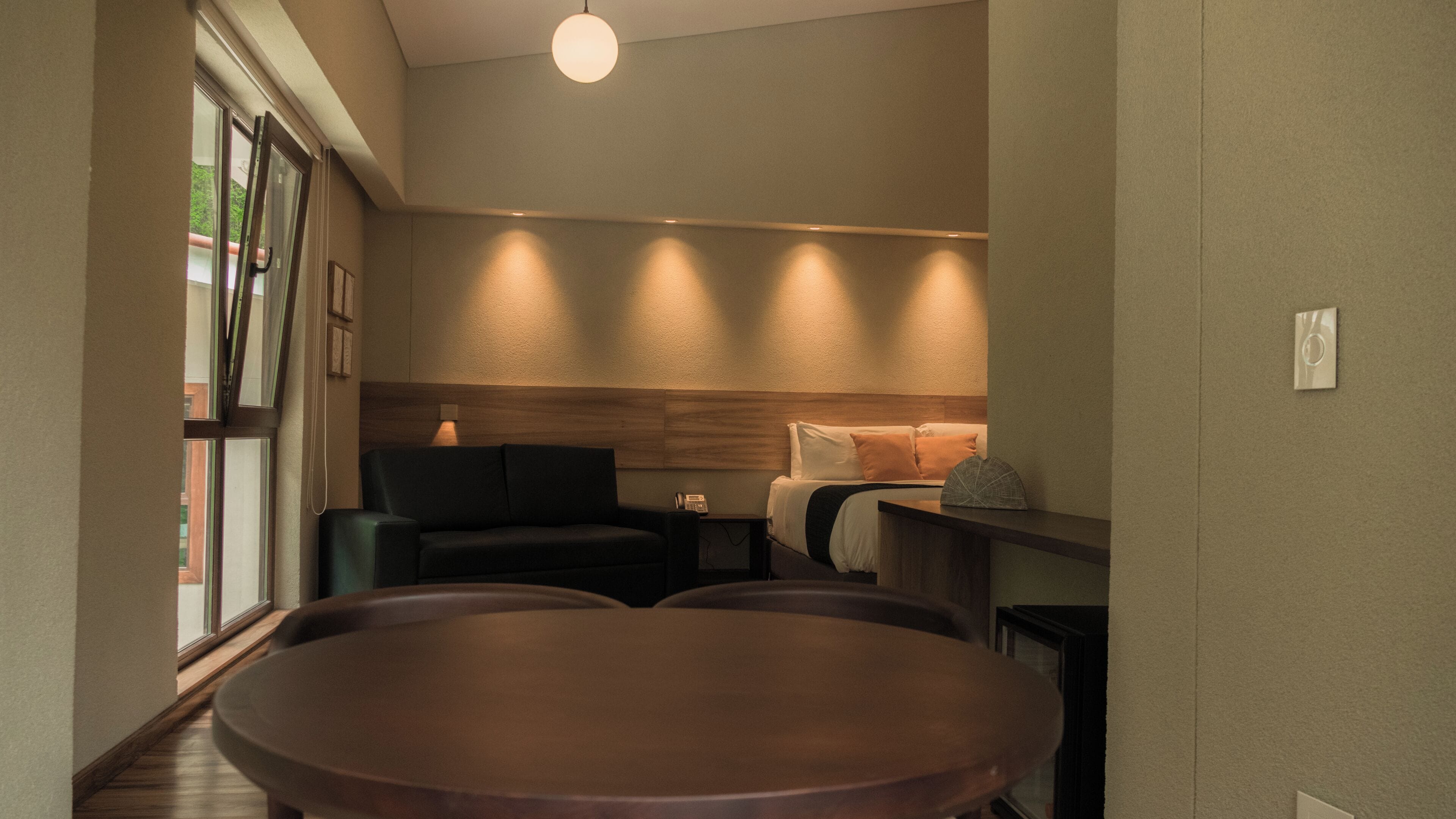 A modern hotel room with a wooden headboard, a sofa, a bed with pillows, a small table, and a large window.