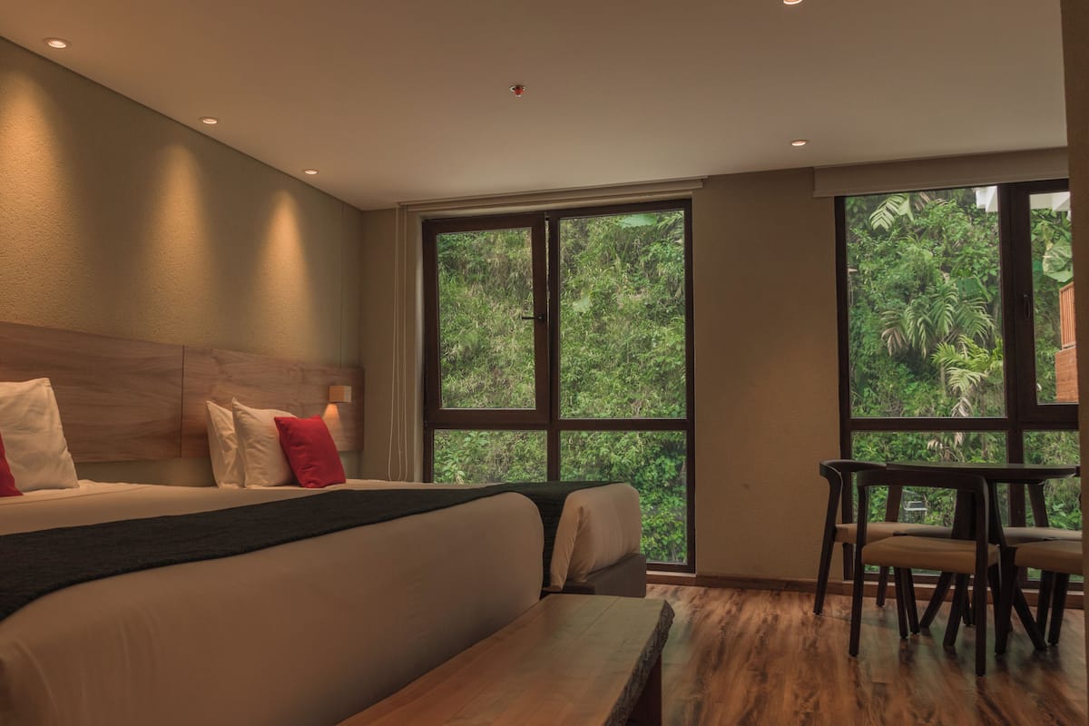Ein modernes Hotelzimmer mit einem großen Bett, einem Holz-Kopfbrett, einem Esstisch und Blick auf üppiges Grün durch große Fenster.