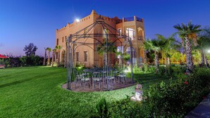 A gazebo with a glass roof surrounded by a well-manicured lawn and palm trees, set against a large building with arched windows and balconies. - La Terra Rosa (Ghmate)