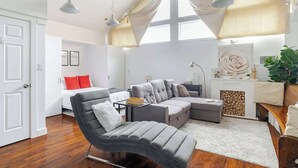 Ein modernes Wohnzimmer mit einer grauen Eckcouch, einem kleinen Tisch, einem roten Zierkissen und einem großen Fenster mit transparenten Vorhängen.