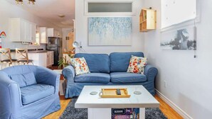 Una sala de estar con un sofá azul, una mesa de centro blanca y una cocina al fondo.