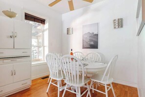 A dining area with a white table and chairs, a window with blinds, and a wall-mounted light fixture.