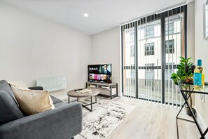 Uma sala de estar moderna com um sofá cinza, uma mesa de centro, uma TV exibindo um filme e uma janela grande com vista para edifícios.