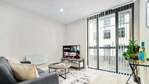 A modern living room with a grey sofa, a coffee table, a TV displaying a movie, and a balcony with a view of buildings. - Luxury 2 Bed Apartment - Smart TV - WIFI (Birmingham)