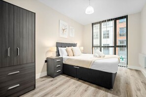 A modern bedroom with a large wardrobe, a bed with a grey headboard, bedside tables with lamps, and a view of a building through large windows. - Stylish 2 Bed Apartment - Smart TV - WIFI -  Jewellery Quarter 1 (Birmingham)