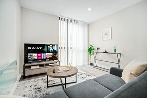 A modern living room with a flat-screen TV displaying a streaming service interface, a grey sofa, a coffee table with a magazine, and a minimalist decor including a plant and a small table with a bottle.