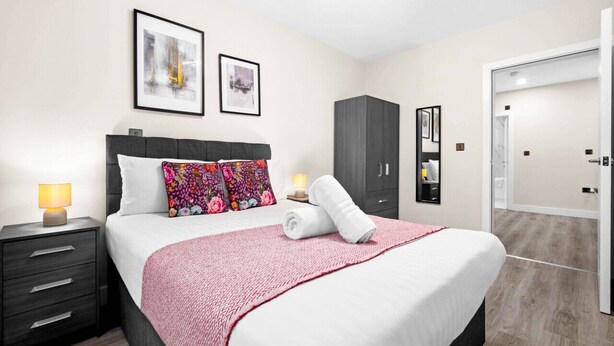 A modern hotel room with a neatly made bed, a nightstand with a lamp, a wardrobe, and a large mirror. - Luxury 2 Bed Apartment - Smart TV - WIFI -  Jewellery Quarter (Birmingham)