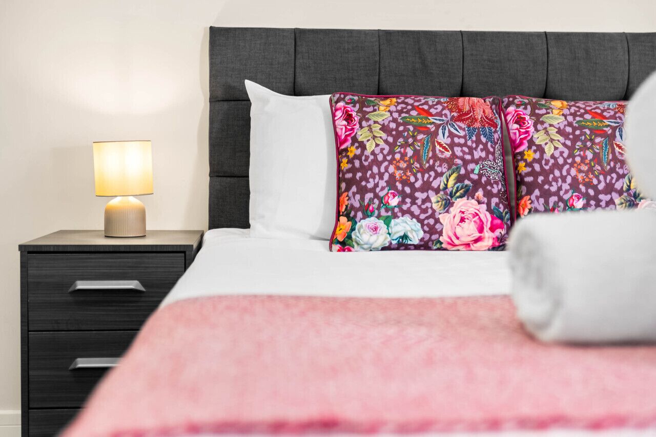 Een net opgemaakt bed met een bloemenkussen, een bedlampje en een nachtkastje in donkere kleuren met lades.