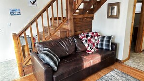 A brown leather sofa with a plaid blanket and pillows, a wooden staircase, and a framed picture on the wall.