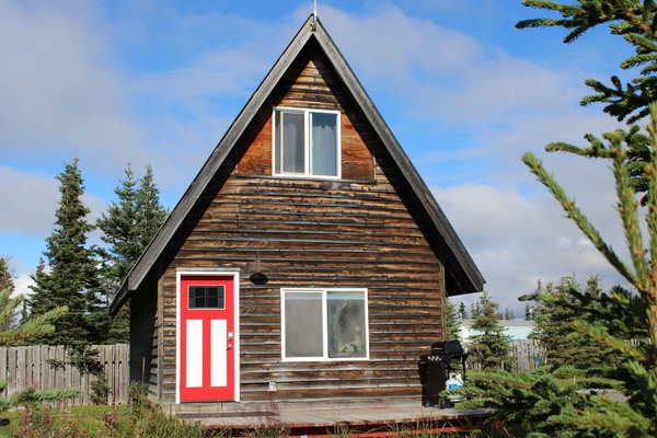 Lush foliage frames this Alaskan cabin, with red door and BBQ for fresh catch.