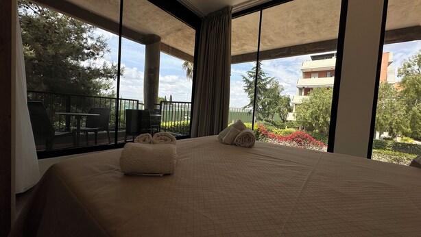 Ein Hotelzimmer mit einem großen Bett, zwei Handtüchern, einem Balkon mit Sitzgelegenheiten und Blick auf Grünflächen sowie Gebäude.