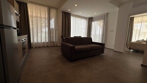Ein modernes Wohnzimmer mit einer Couch, einer Küchenzeile mit Kühlschrank und großen Fenstern mit Vorhängen.