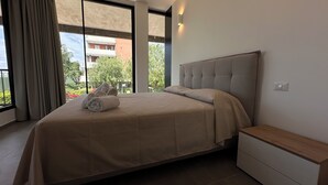 Ein modernes Schlafzimmer mit einem großen Bett, einem Nachttisch und Blick auf ein Gebäude durch ein Fenster.