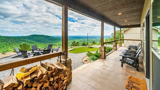 Un espace extérieur abrité, doté de sièges, d’une pile de bois de chauffage et offrant une vue sur un paysage verdoyant.