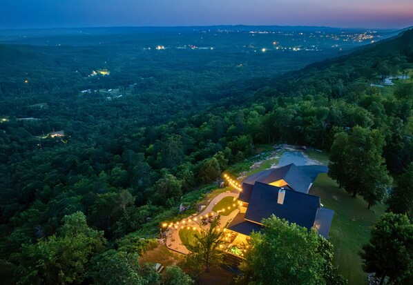 A house with a dark roof, surrounded by dense forest, and a string of lights along the driveway.