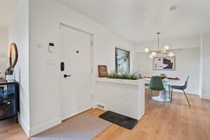 A modern hotel room with a wooden floor, a white door, a small desk with a mirror, a bedside table, and a bed. There is a dining area with a table and chairs, a chandelier, and a framed picture on the wall. - Yard w/ fire pit & grill, king bed, light filled (Boise)