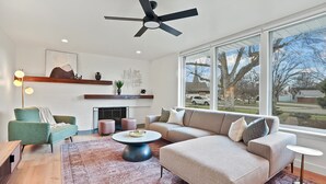 A modern living room with a fireplace, a large window with a view, a ceiling fan, and a coffee table. - Yard w/ fire pit & grill, king bed, light filled (Boise)